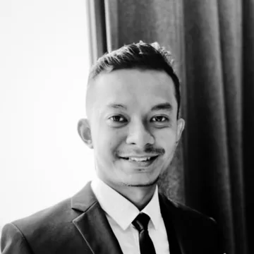 Azwan Zuharimi, in a black and white photograph, smiling in a dark suit and tie beside a curtain.
