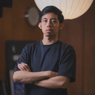 Hazqeel Afyq, in a navy T-shirt with his arms crossed, under a paper lamp in a wood-paneled room.