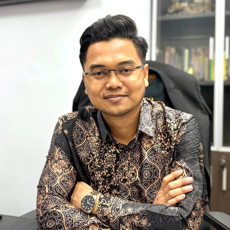 Afiq Asri, in rimless glasses and a black and gold batik shirt, smiling slightly with his arms crossed at a desk.