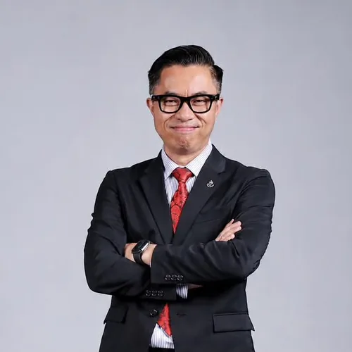 Ahmad Faisal Mohamad Ayob, smiling with his arms folded, in a dark suit, light shirt and red tie against a grey background.