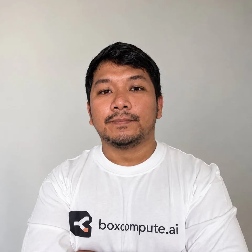 Farhan Helmy, with a slight smile, in a white boxcompute.ai T-shirt against a light gray wall.