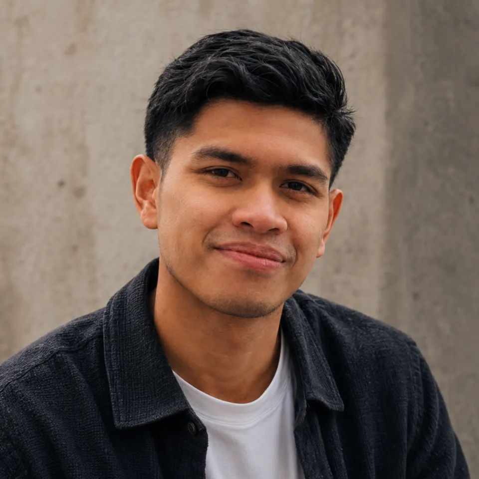 Syahmi Rafsanjani Shaarani, smiling slightly, in a dark textured overshirt and white T-shirt in front of a concrete wall.